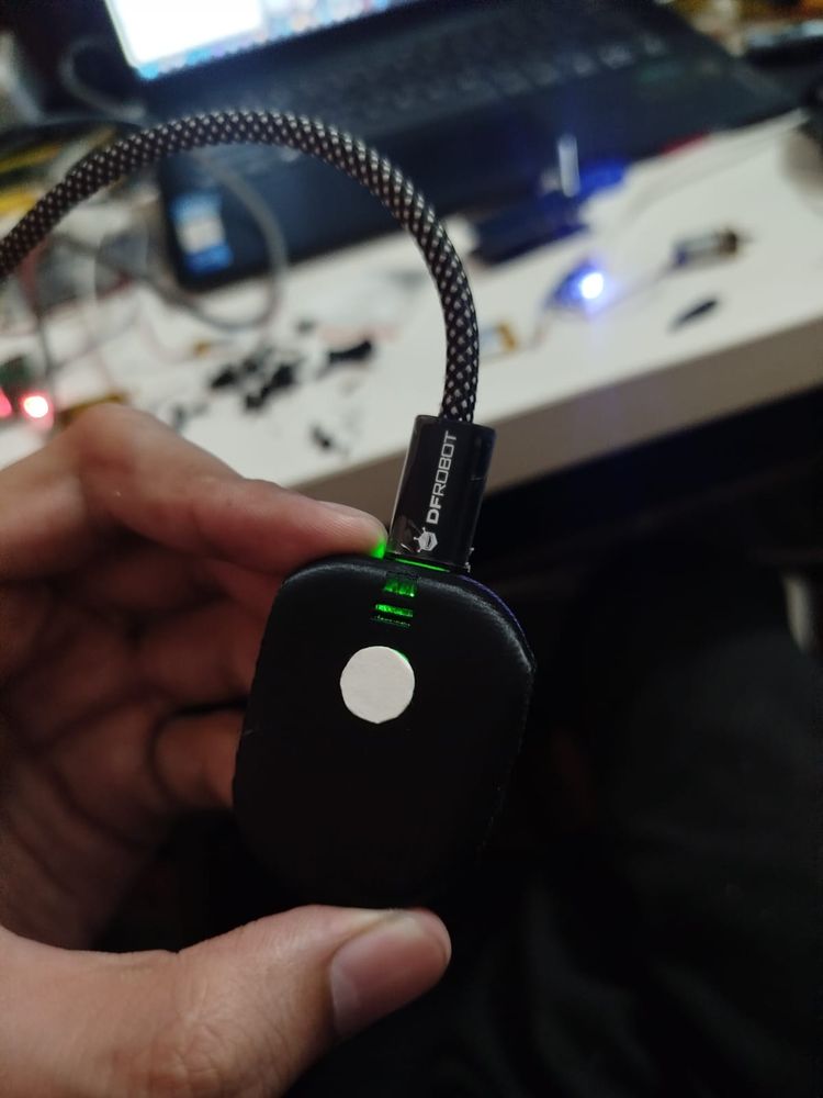Pebble-shaped wearable on its USB charging cable with status LED lit, held in hand